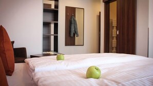 A hotel room with a bed, two green apples on the bed, a coat hanging on the wall, and a wooden wardrobe.