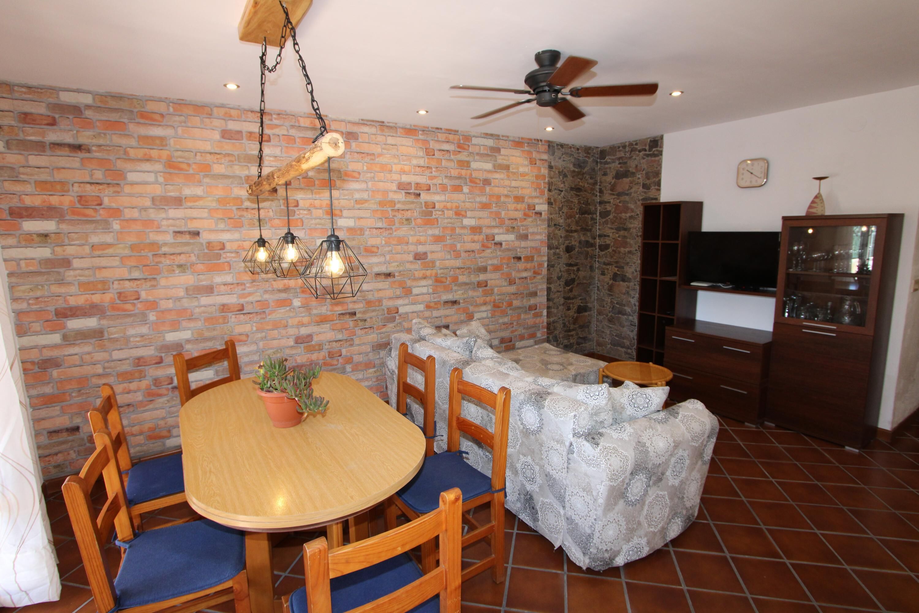 A dining area with a brick wall, wooden chairs, a round table, and hanging light fixtures.