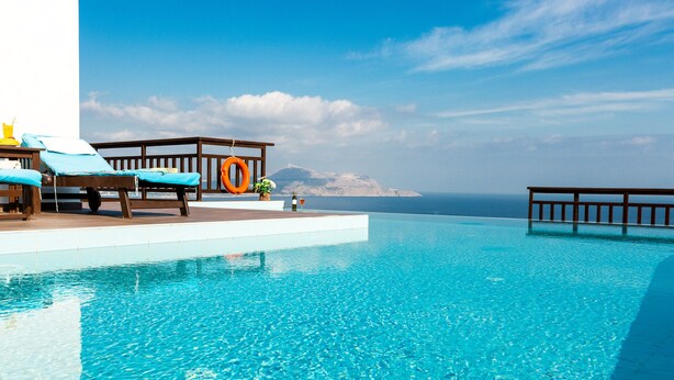 Een helderblauw zwembad met ligbedden en een houten terras met uitzicht op de zee.