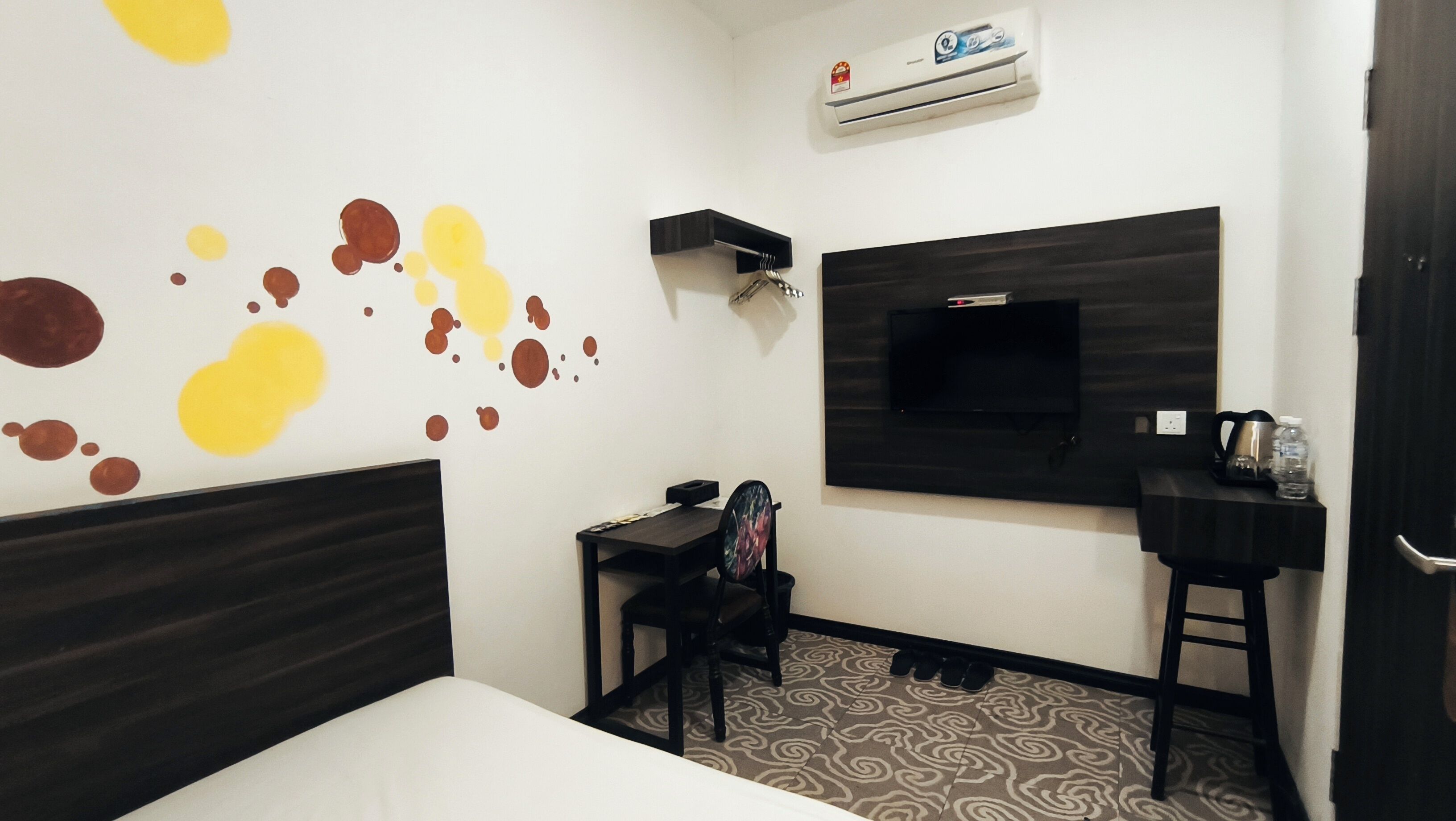 Una habitación de hotel con cama, escritorio, silla, televisor montado en la pared y un zapatero.