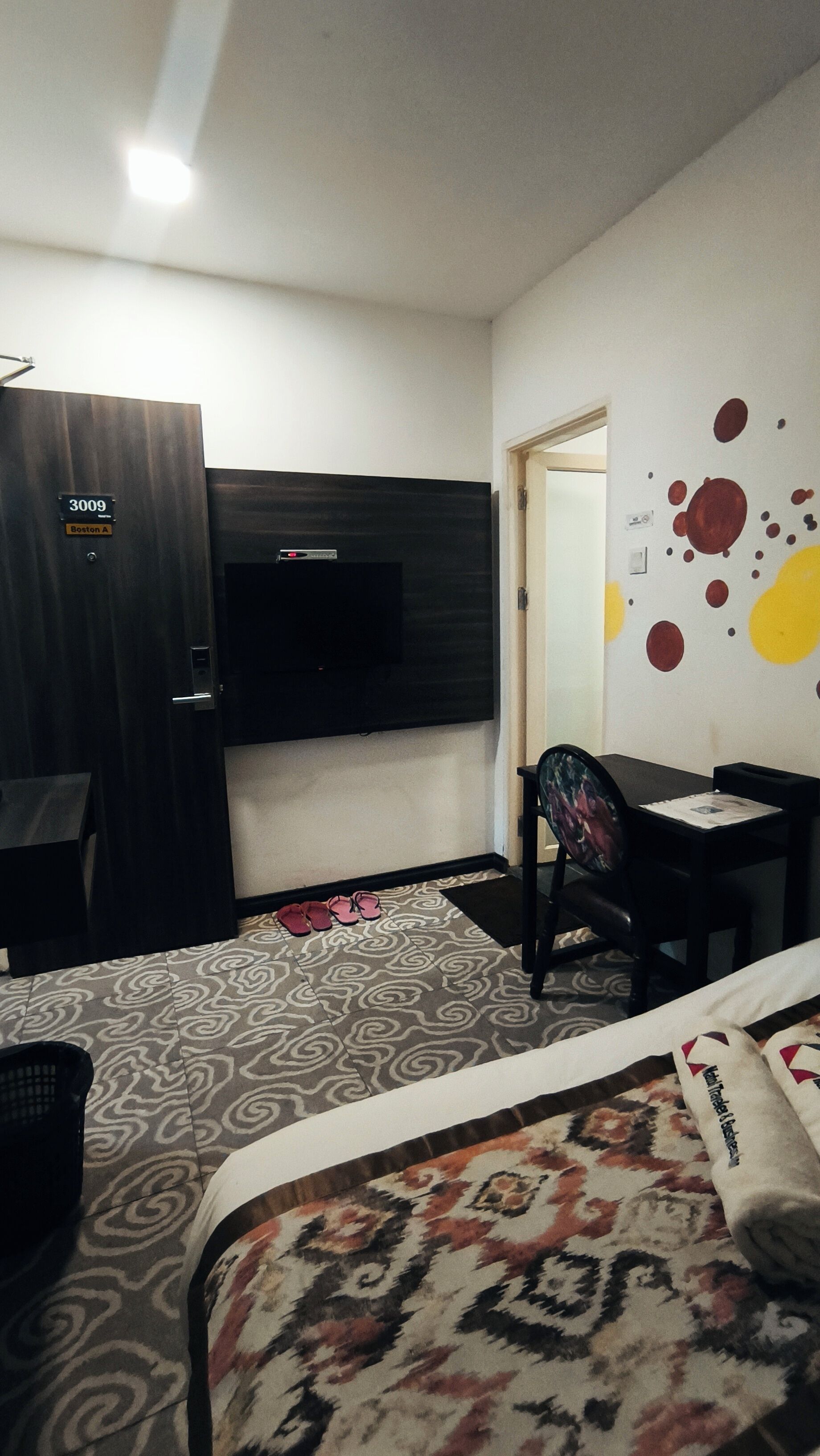 A hotel room with a bed, a desk, a chair, a TV mounted on the wall, and a door with a sign.