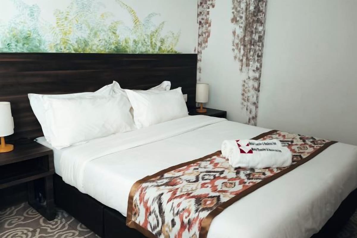 A hotel room with a large bed, white bedding, a patterned throw blanket, and a wall painting.