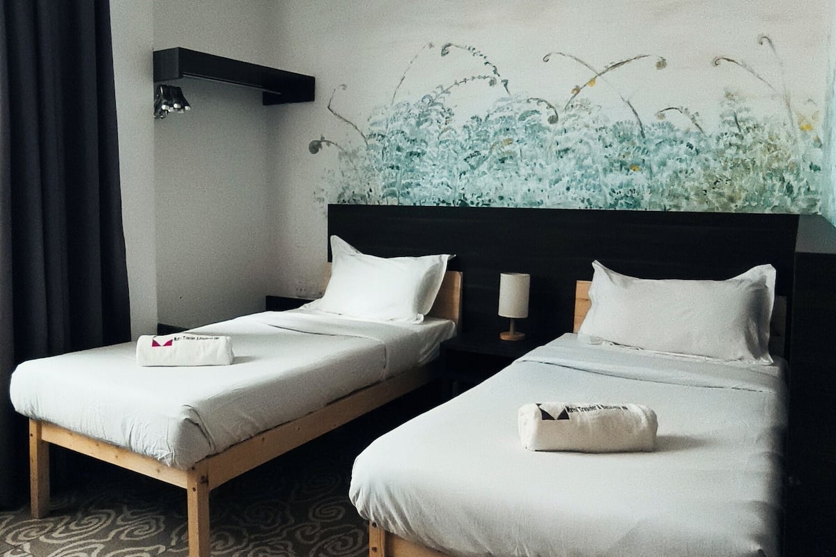 A hotel room with two single beds, a patterned carpet, a wall-mounted air conditioner, and a painting above the beds.