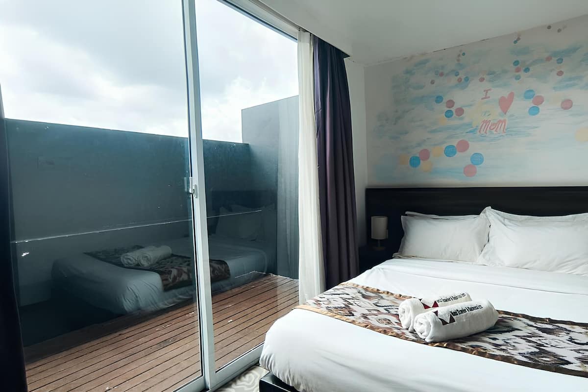 A hotel room with a large bed, a wooden floor, a glass door leading to a balcony, and a colorful wall mural.