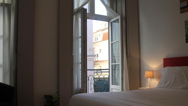 Habitación de hotel con cama, mesita de noche con lámpara, planta en maceta y ventana abierta que da a un balcón y edificios exteriores.