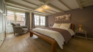 Een slaapkamer met een groot bed, een bankje, een stoel, een nachtkastje en een raam met gordijnen.