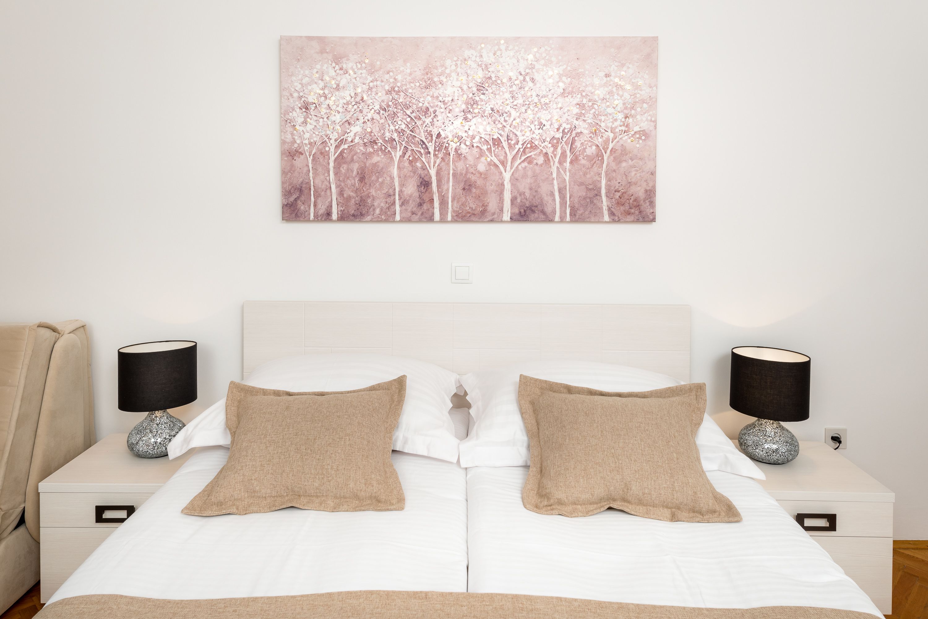 A bedroom with a white bed, two beige pillows, a white headboard, a wall-mounted painting of trees, and two bedside tables with lamps.