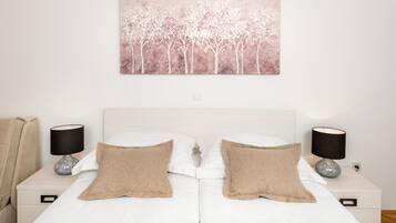 A bedroom with a white bed, two beige pillows, a white headboard, a wall-mounted painting of trees, and two bedside tables with lamps.