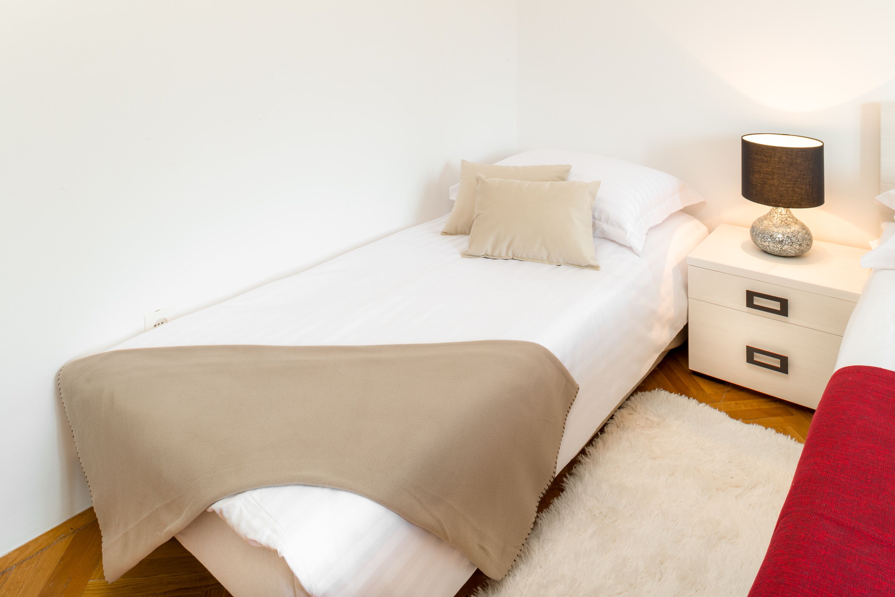 A small, neatly made bed with a beige blanket and pillows, a white nightstand with a lamp, and a red chair.