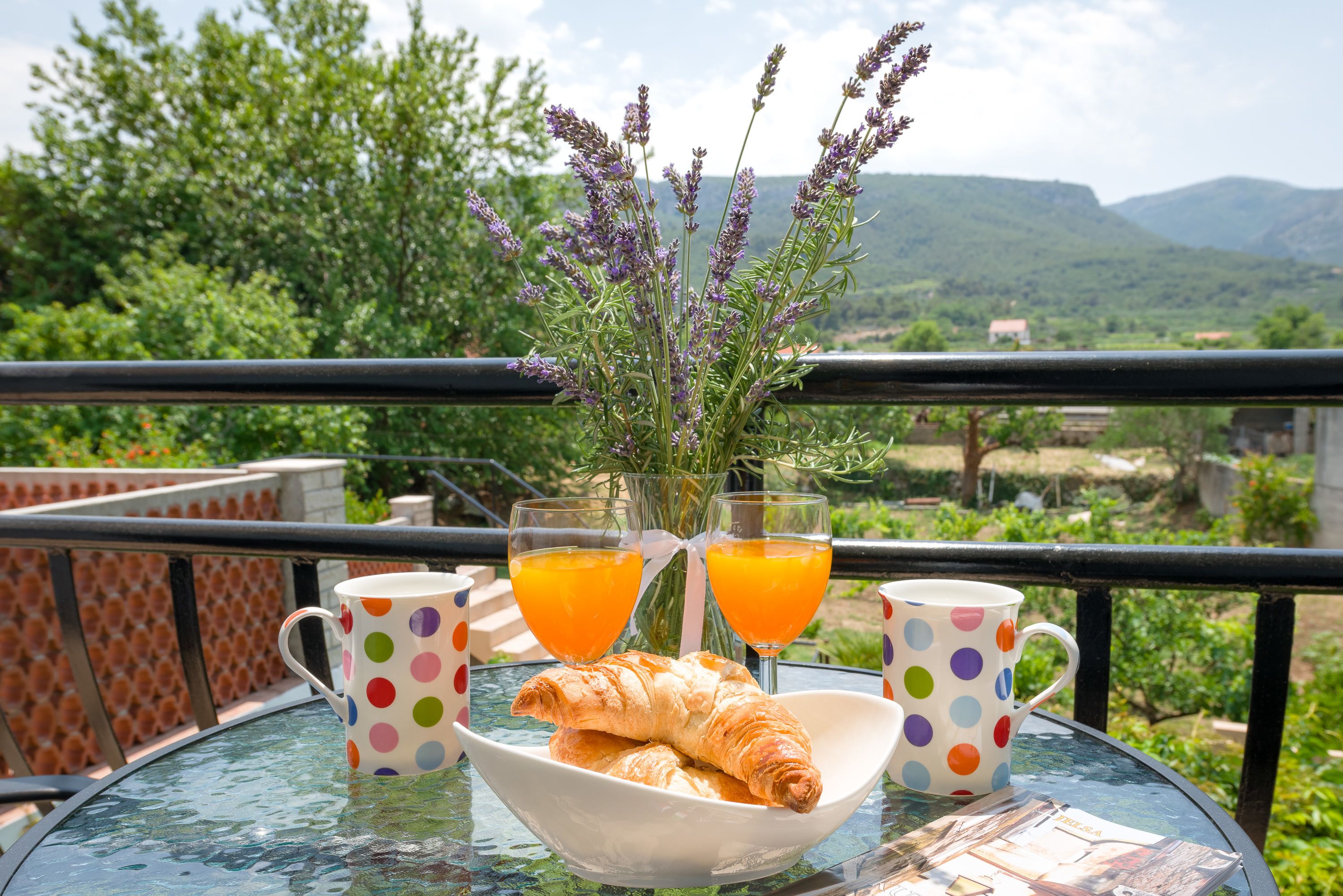 Uma mesa com um recipiente de croissants, dois copos de sumo de laranja e um vaso de lavanda numa varanda com vista para uma paisagem deslumbrante.