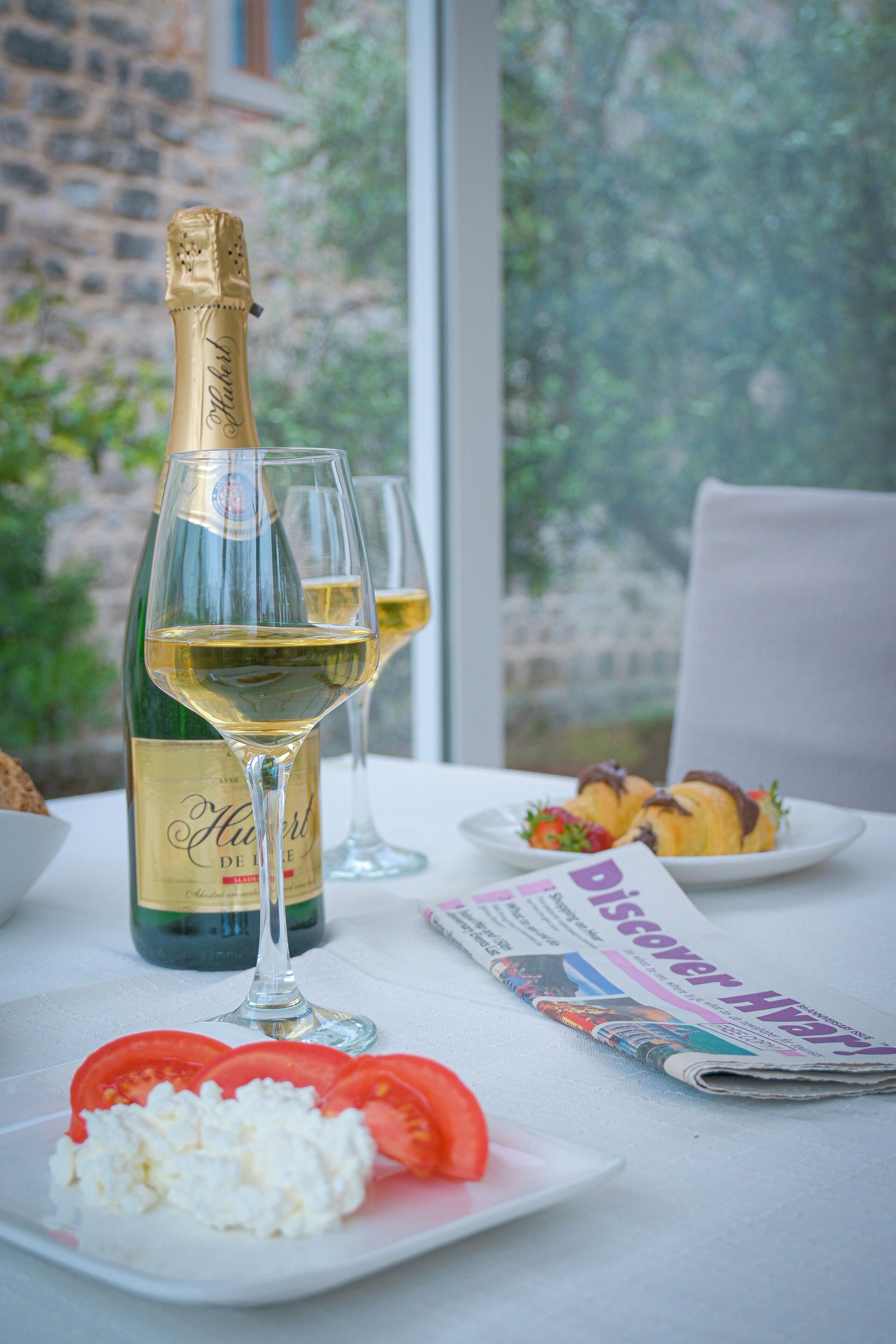 A table with a bottle of wine, two glasses, a plate with tomatoes and cheese, and a folded newspaper.