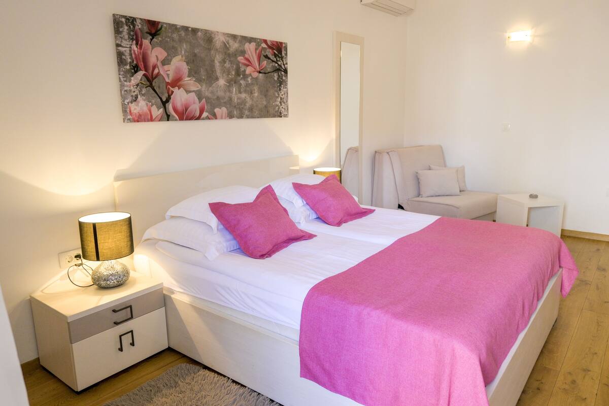 A bedroom with a bed, bedside tables, a nightstand, a lamp, a mirror, and a painting on the wall.