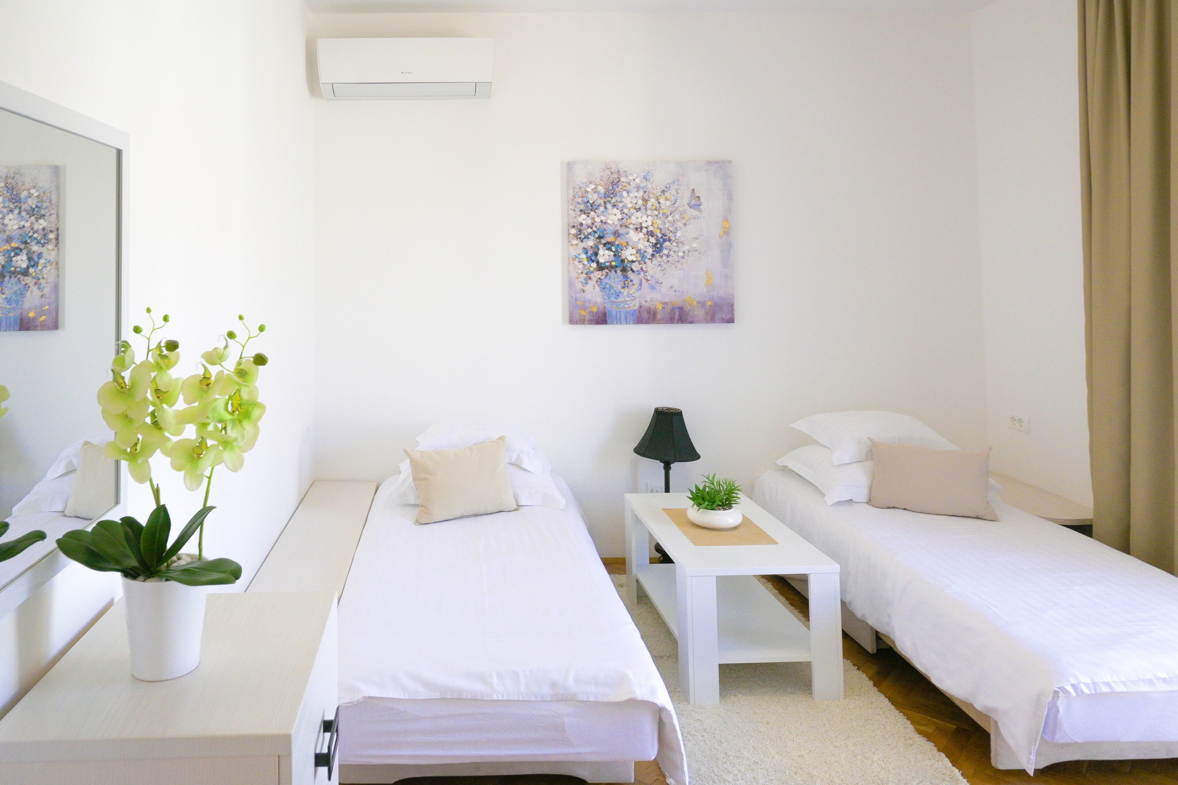 A hotel room with two single beds, a small table with a lamp, a potted plant, and a painting on the wall.