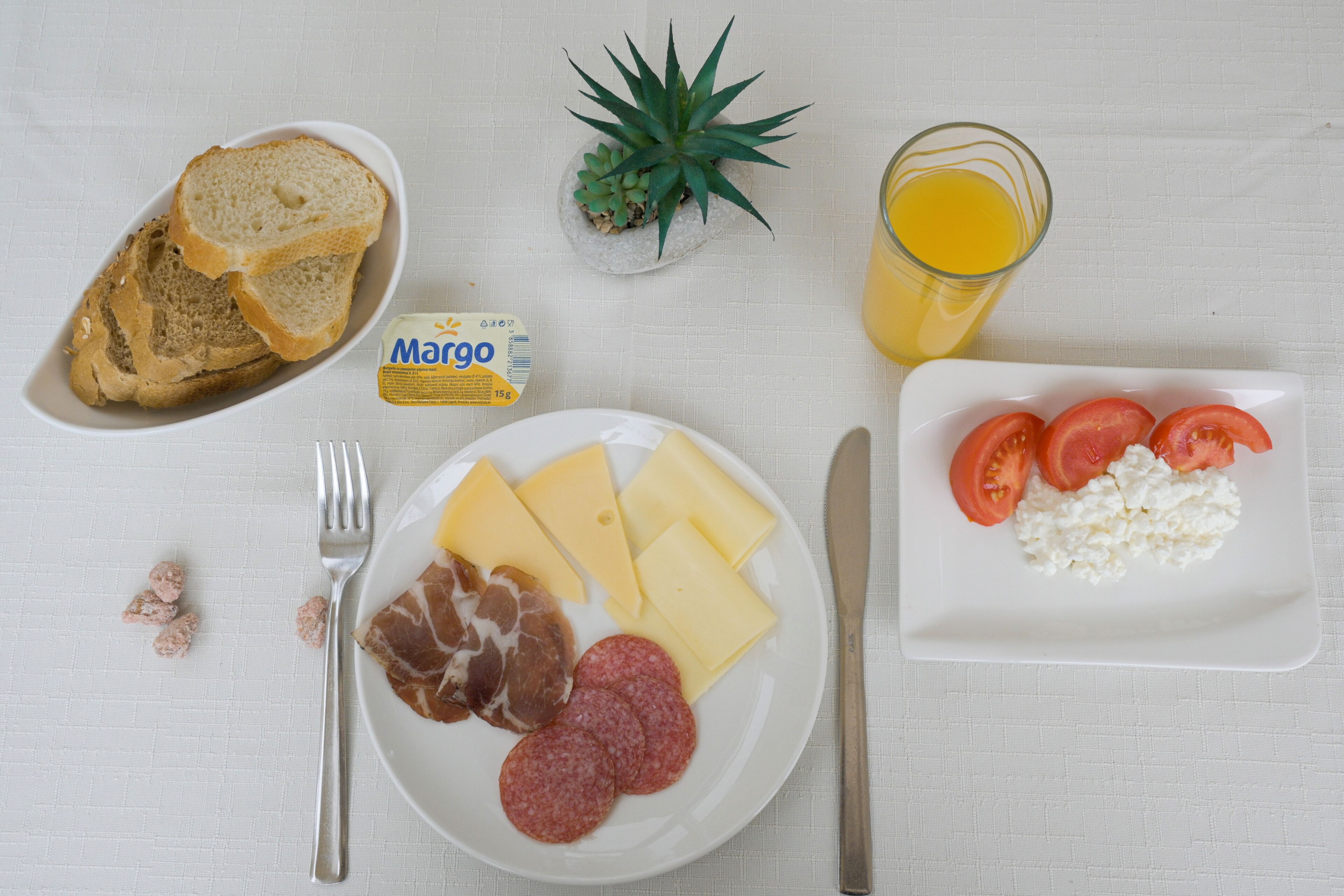 Un piatto per la colazione con pane, formaggio, prosciutto e pomodori, un bicchiere di succo d'arancia e una piccola pianta su una tovaglia bianca.