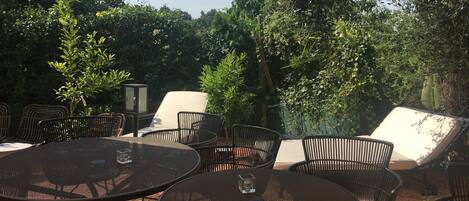 An outdoor patio with round metal tables and wicker chairs, surrounded by dense greenery.