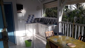 A kitchen with white cabinets, a tiled backsplash, and a small island. There is a dining table with chairs and a green trash bin. - ti case gwada bungalow rentals "ti dauphin" (Sainte-Anne)
