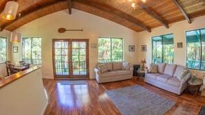 A spacious living room with a high wooden ceiling, a beige sofa, a dining area with a wooden table, and large windows offering a view of the outdoors. - 6 mins from Nat'l Park! Enjoy the sound of rain & birds in a peaceful retreat. (Volcano)