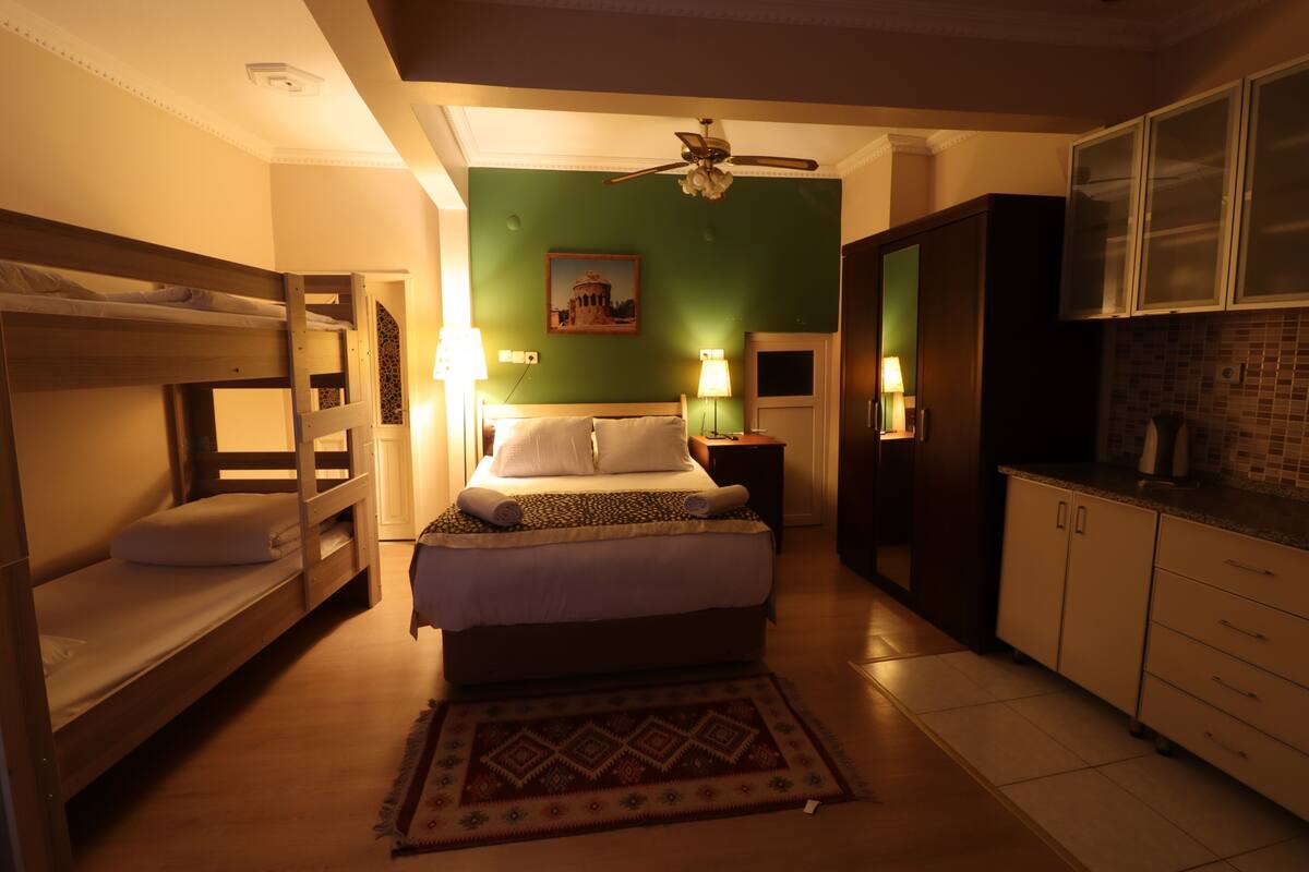 A compact hotel room with a bunk bed, a single bed, a ceiling fan, a kitchen area with cabinets, and a patterned rug.