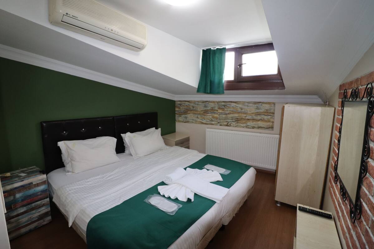 A hotel room with a bed, a green bedspread, white bedding, a radiator, a window with curtains, a wardrobe, and a mirror on the wall.