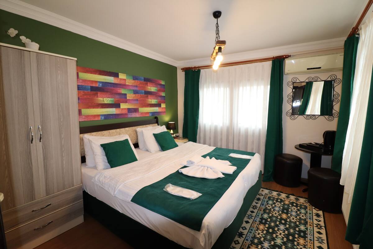 A neatly made bed with green bedding, a colorful abstract wall art, a wooden wardrobe, and a small round table with a lamp.