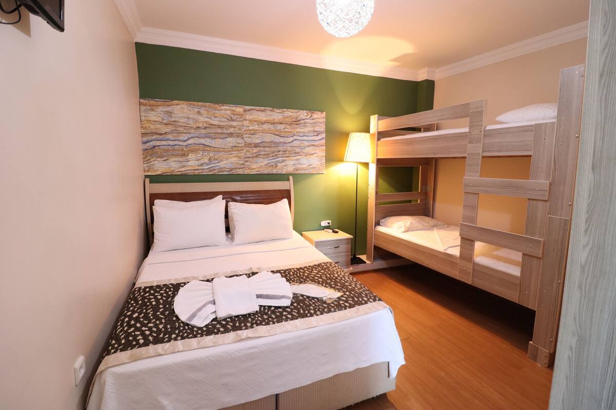 A hotel room with a large bed, a smaller bunk bed, a bedside table with a lamp, and a wall art piece.