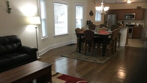 Un salon avec un canapé en cuir foncé, une table basse en bois et un tapis rouge et beige. On y trouve également un coin repas avec une table et des chaises en bois, ainsi qu’une cuisine équipée d’un four à micro-ondes et d’un four traditionnel.