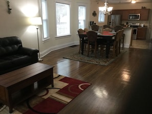 Una sala de estar con un sofá de cuero oscuro, una mesa de centro de madera y un tapiz rojo y beige. Hay una zona de comedor con una mesa y sillas de madera, y una cocina con microondas y horno.