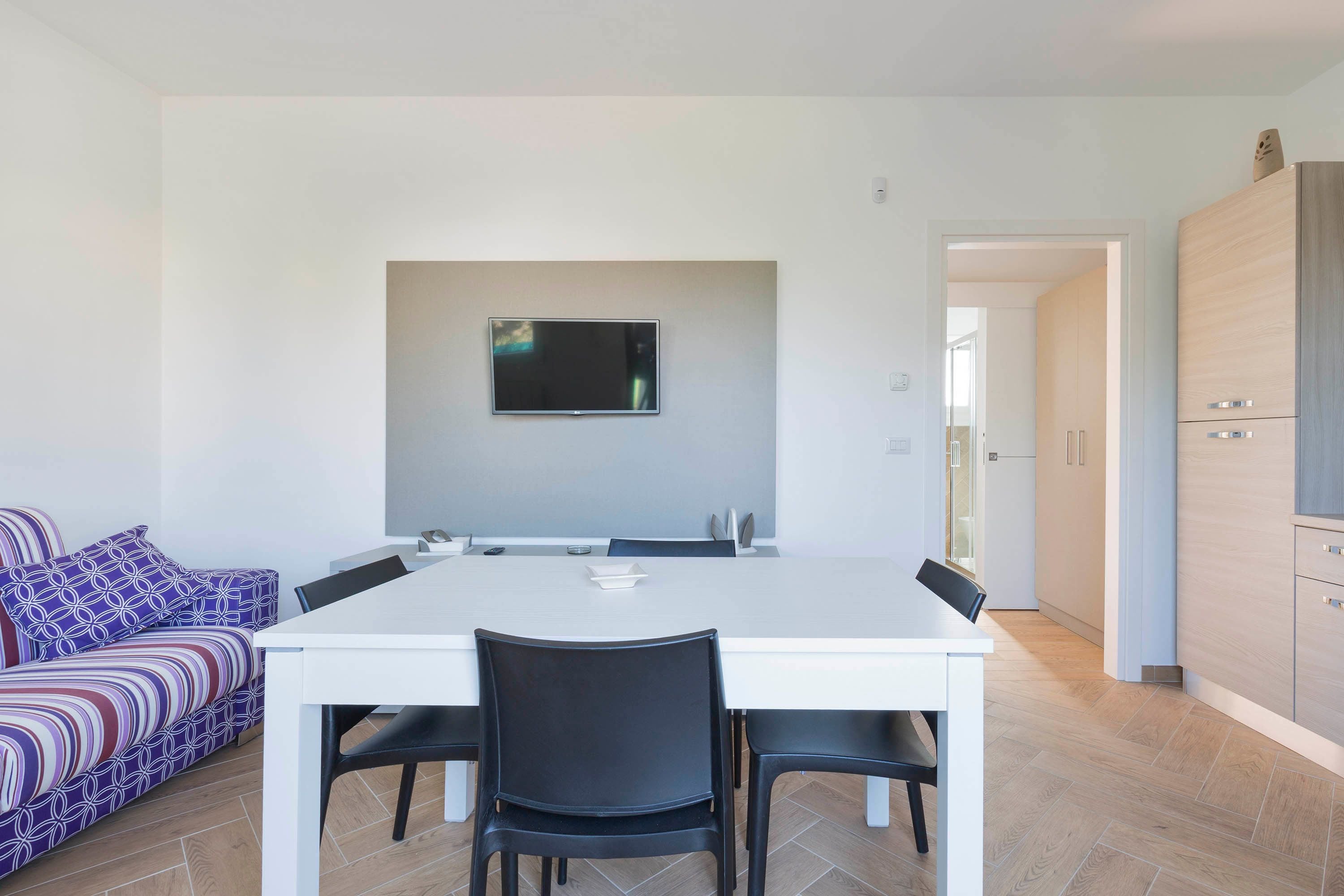 Una cocina moderna con una mesa y sillas blancas, un televisor de pantalla plana montado en la pared, y un sofá estampado en el área de estar contigua.