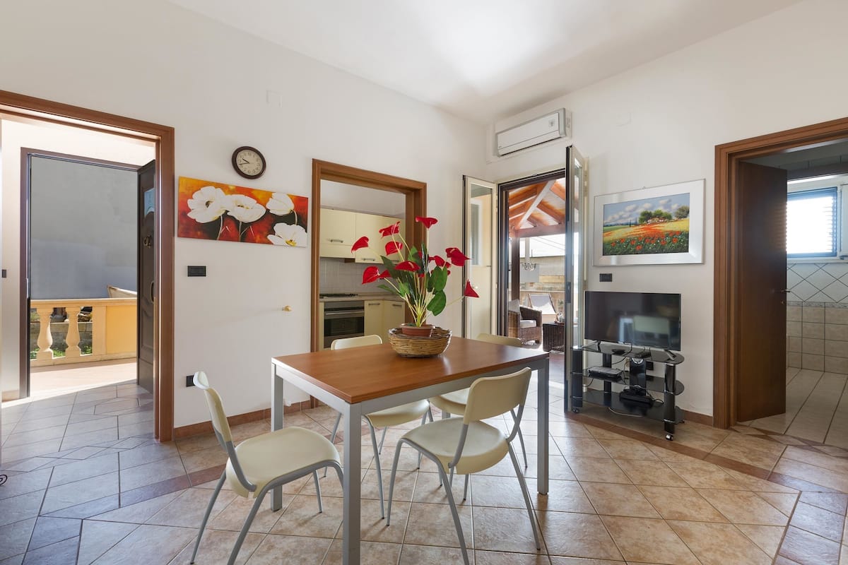 A dining area with a wooden table, four chairs, a potted plant, and a television on a stand.