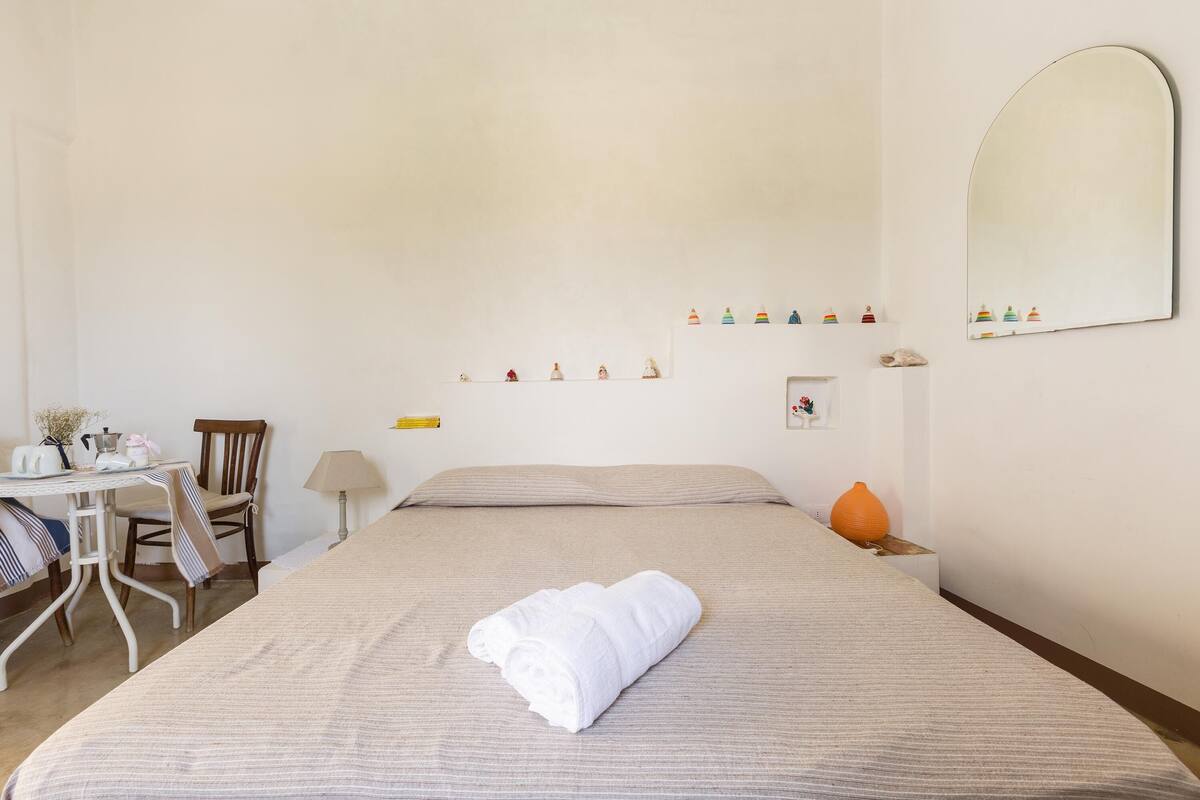 A neatly made bed with a rolled white towel, a small table with a lamp, a chair, and a shelf with decorative items.