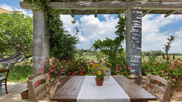 Una mesa de madera al aire libre con mantel blanco, rodeada de sillas, bajo una pérgola con vistas a un jardín lleno de plantas y flores.
