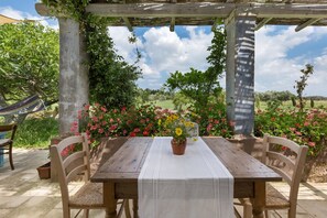 Mesa de madeira ao ar livre com toalha branca, rodeada por cadeiras, sob uma pérgula com vista para um jardim com diversas plantas e flores.
