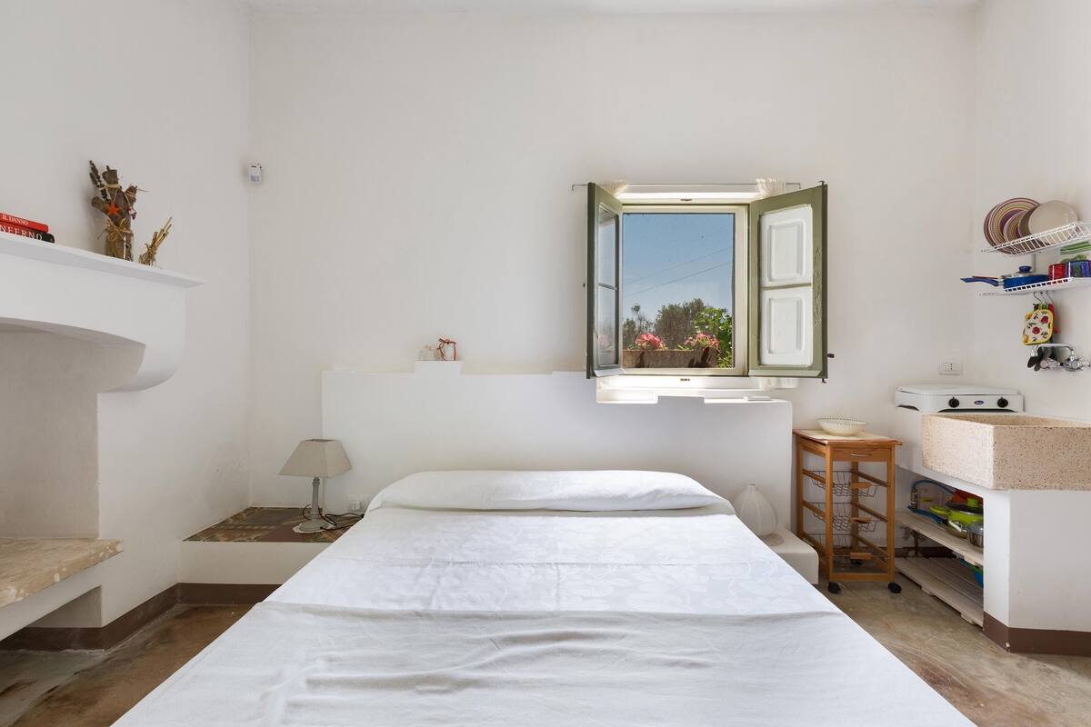 A minimalist bedroom with a bed, a bedside table, a lamp, a small wooden stool, and a window with a view of greenery.