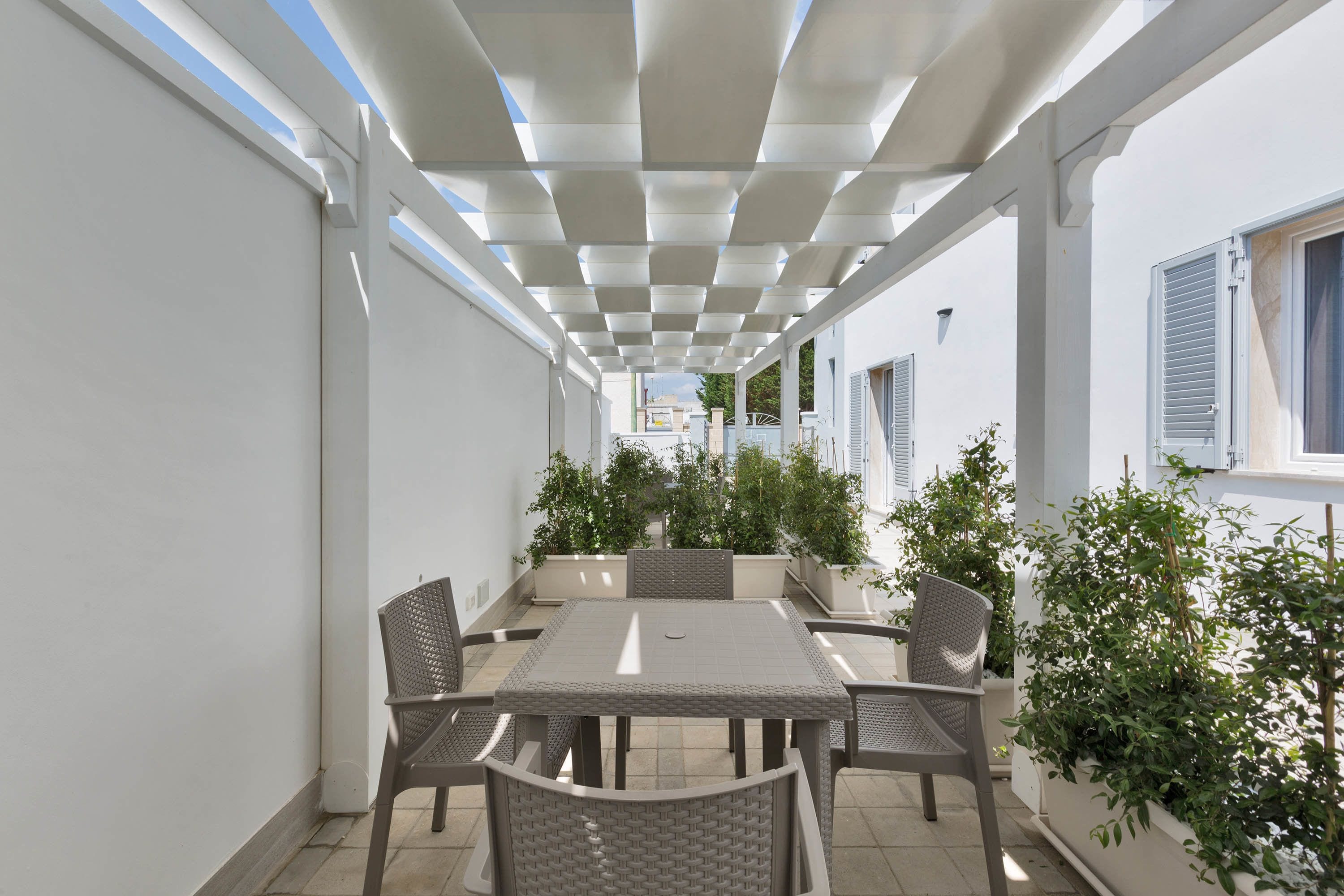 A covered outdoor patio with a table and chairs, surrounded by potted plants and white walls.
