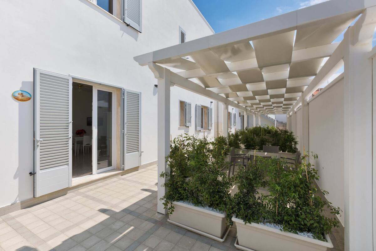 A covered outdoor area with a pergola, potted plants, and a white building with shutters.