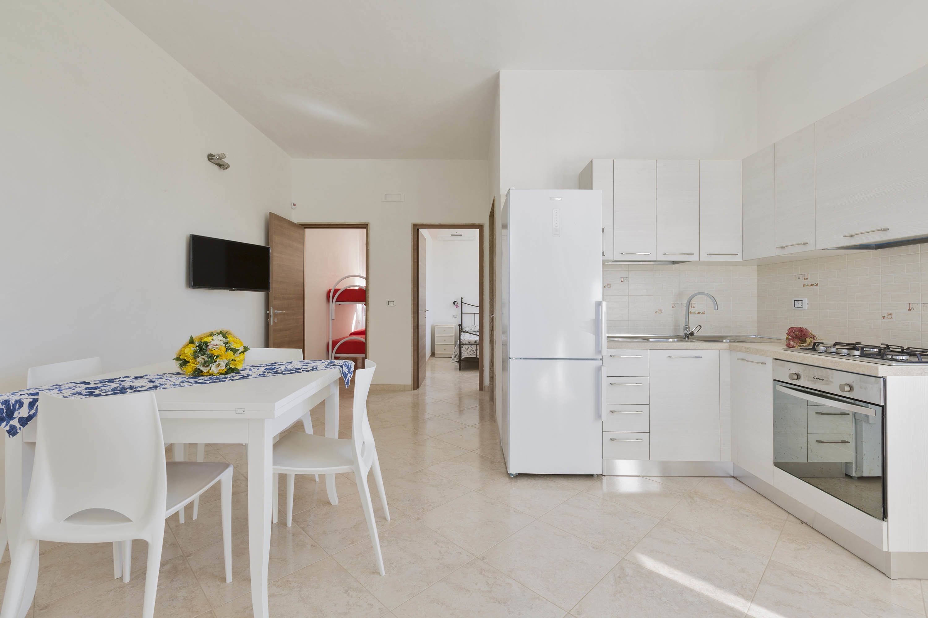 A modern kitchen with white cabinets, a refrigerator, and an oven. There is a dining table with chairs and a TV mounted on the wall.