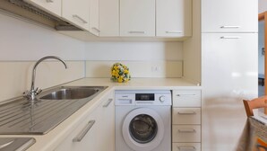 A compact kitchen with a built-in washing machine, a stainless steel sink, and white cabinetry. - Residence Tabù (Porto Cesareo)