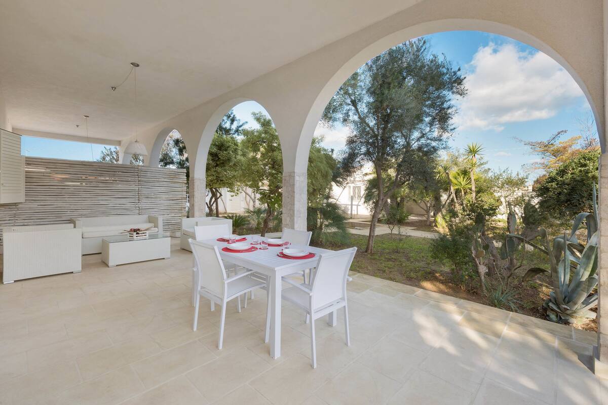 An outdoor dining area with white furniture and red tableware, surrounded by arches and a view of trees and plants.