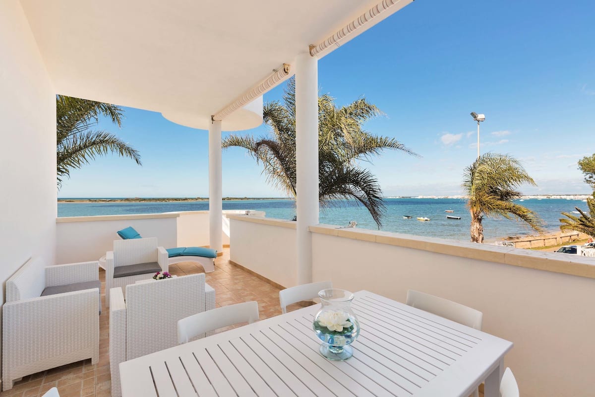 A balcony with white wicker furniture, a glass vase with flowers, and a view of the sea and palm trees.