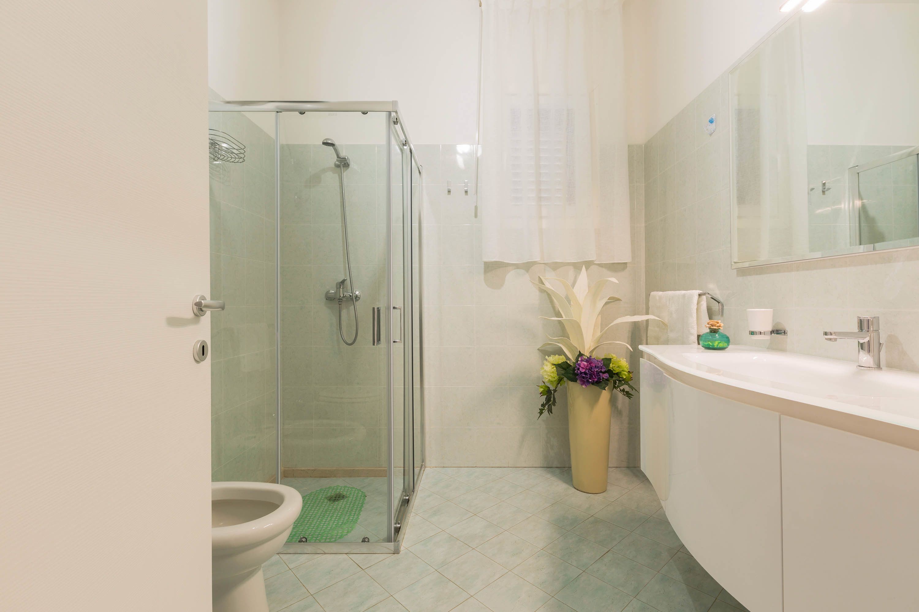 Un baño moderno con ducha acristalada, inodoro, lavamanos con espejo y un florero con flores.
