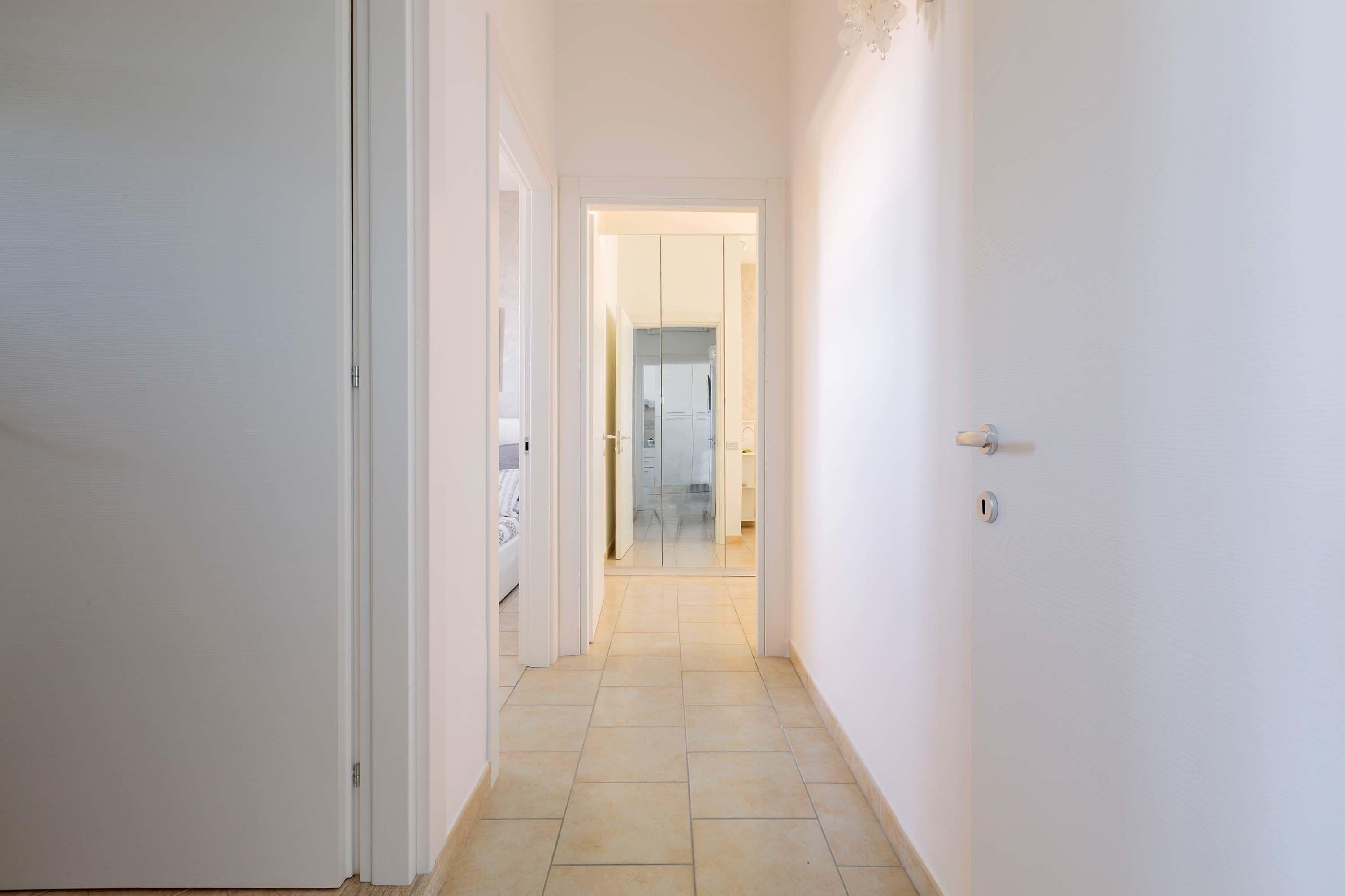 A narrow hallway with white walls and a tiled floor leading to a room with a glass door.