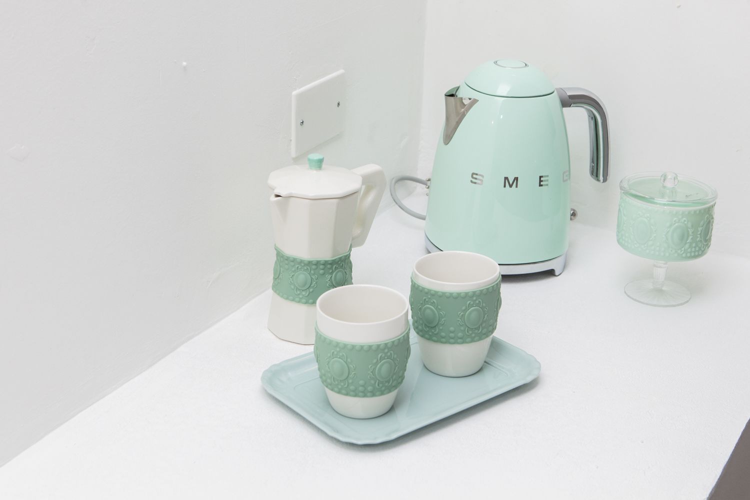 A kitchen setup with a white countertop, a white kettle, and matching cups and a glass on a tray.