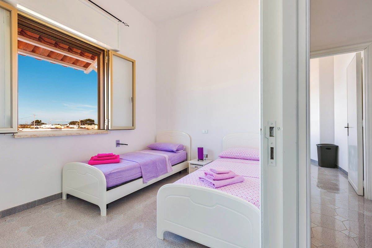 A bedroom with two single beds, a window with a view, a small bedside table, and a trash bin.