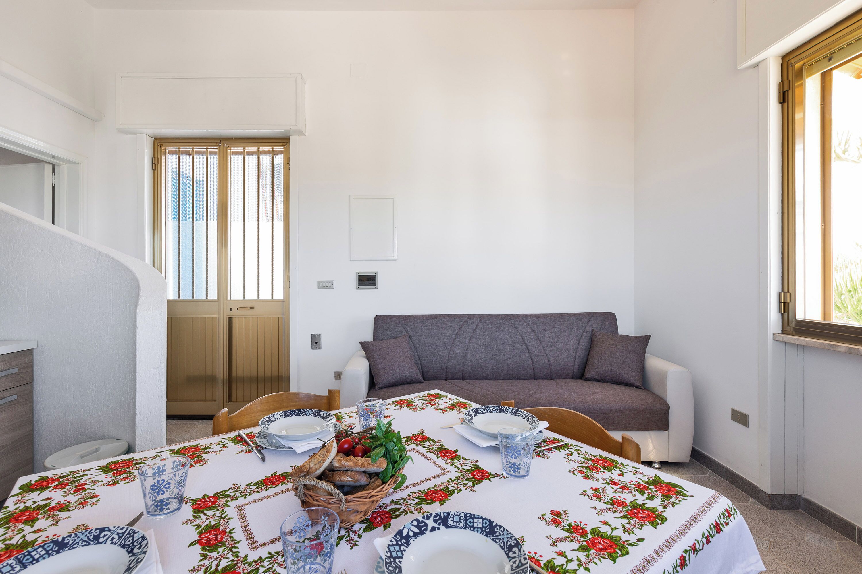 Un coin repas avec une table dressée pour quatre personnes, un canapé gris et une fenêtre avec des stores.