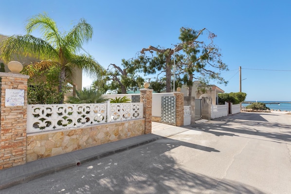 A gated entrance with stone walls and decorative metalwork, palm trees, and a view of the sea.