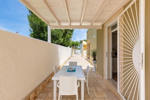 A covered outdoor patio with a white table and chairs, a potted plant, and a view of the street and buildings in the background.