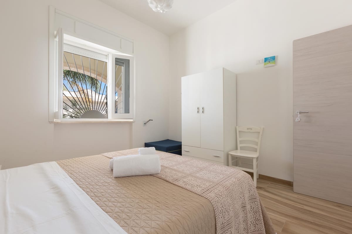 A bedroom with a bed, a wardrobe, a chair, and a window with a view of palm trees.