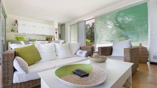 A modern living room with a white sofa, green and white woven coffee table, wicker side tables, and a view of the outdoors through large windows. - Villa Carlita (Carovigno)