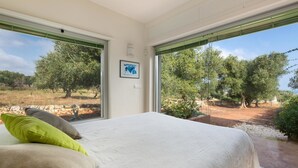A bedroom with a large bed, two pillows, a framed picture on the wall, and a view of trees and a stone wall outside. - Villa Carlita (Carovigno)