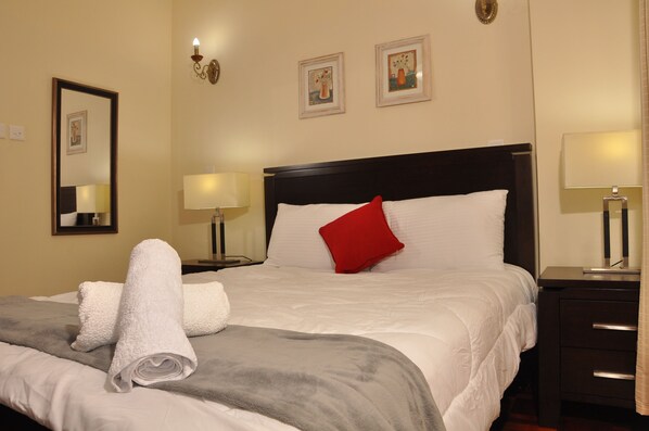A neatly made bed with white linens and a red pillow, a grey blanket, and two rolled white towels on a wooden headboard.